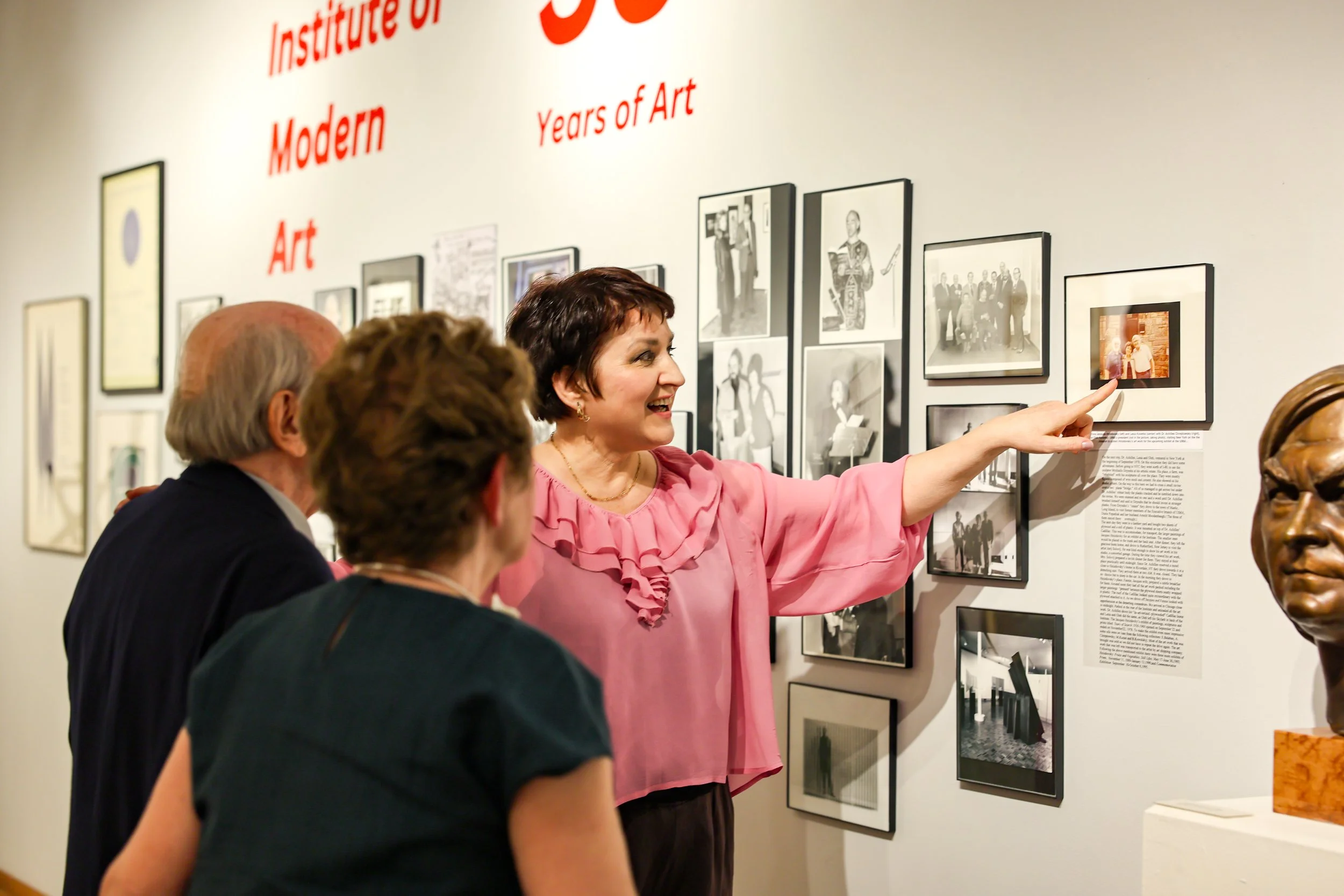 Elena Smolynets pointing out photographs on a wall retrospective celebrating fifty-five years of the Institute