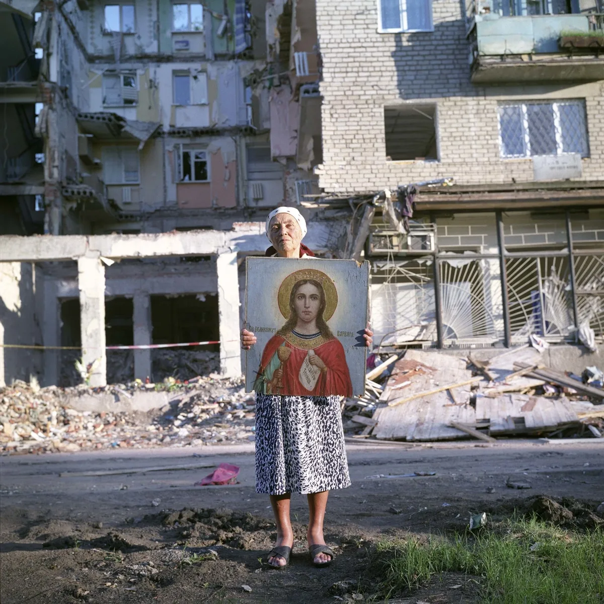 An elderly woman standing in front of a bombed-out apartment block, holding a religious icon
