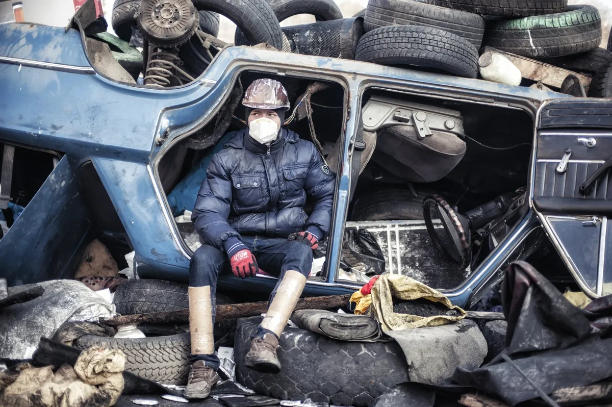 Photograph of a masked protester sitting inside an overturned car among stacked tires on a barricade